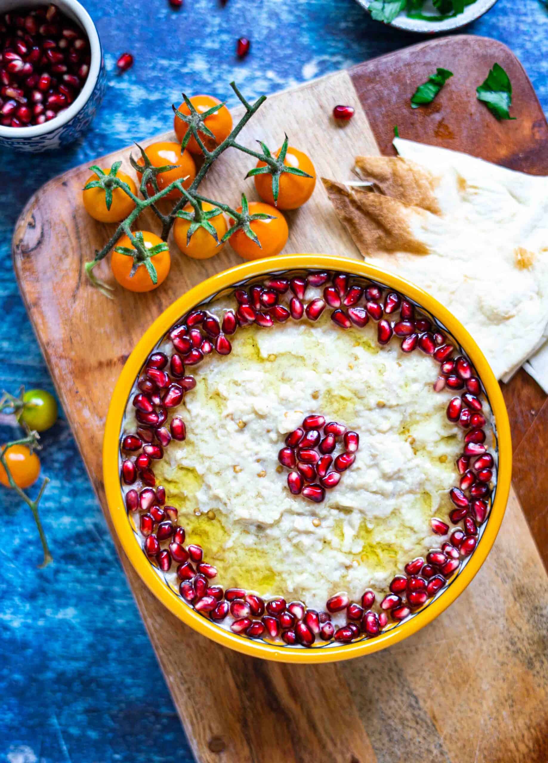 mutabal on plate decorated with pomegranate seeds and extra virgin olive oil, next to bread and grapes tomatoes.