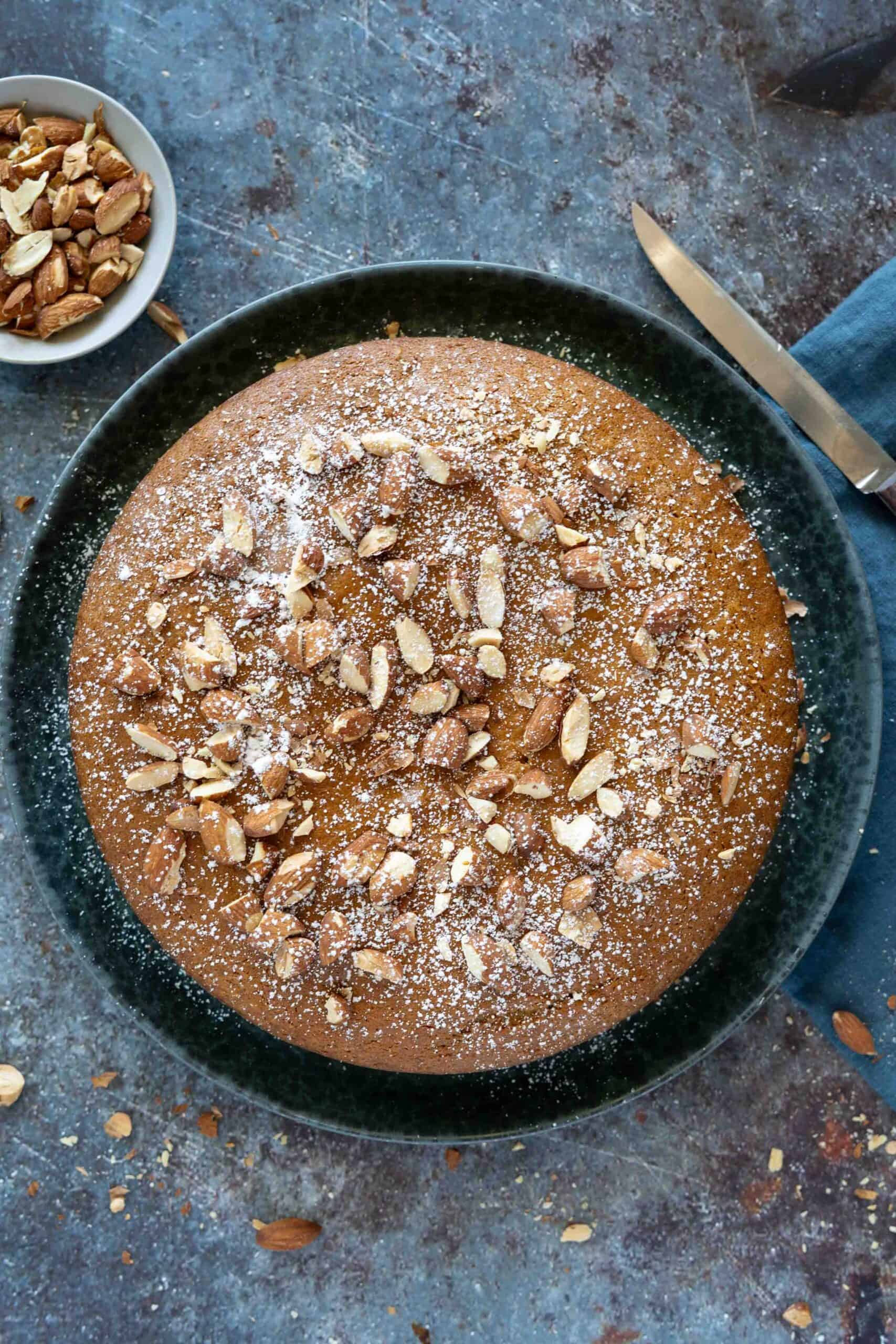 The cake is served on a large plate, garnished with toasted chopped almonds and a dusting of powdered sugar.