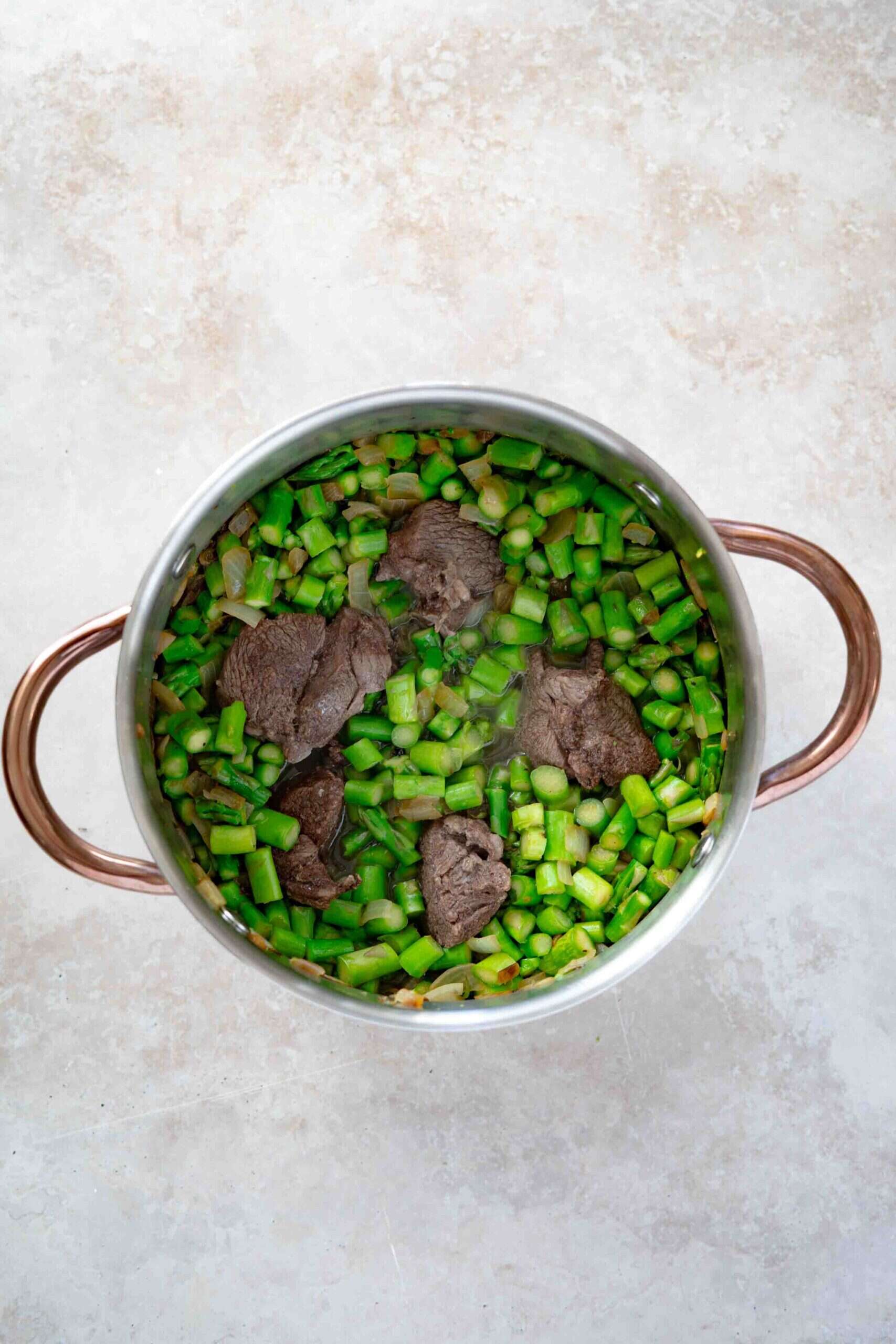 Pot filled with sautéed onions and chopped asparagus mixed with sauteed onions topped with tender lamb and broth.