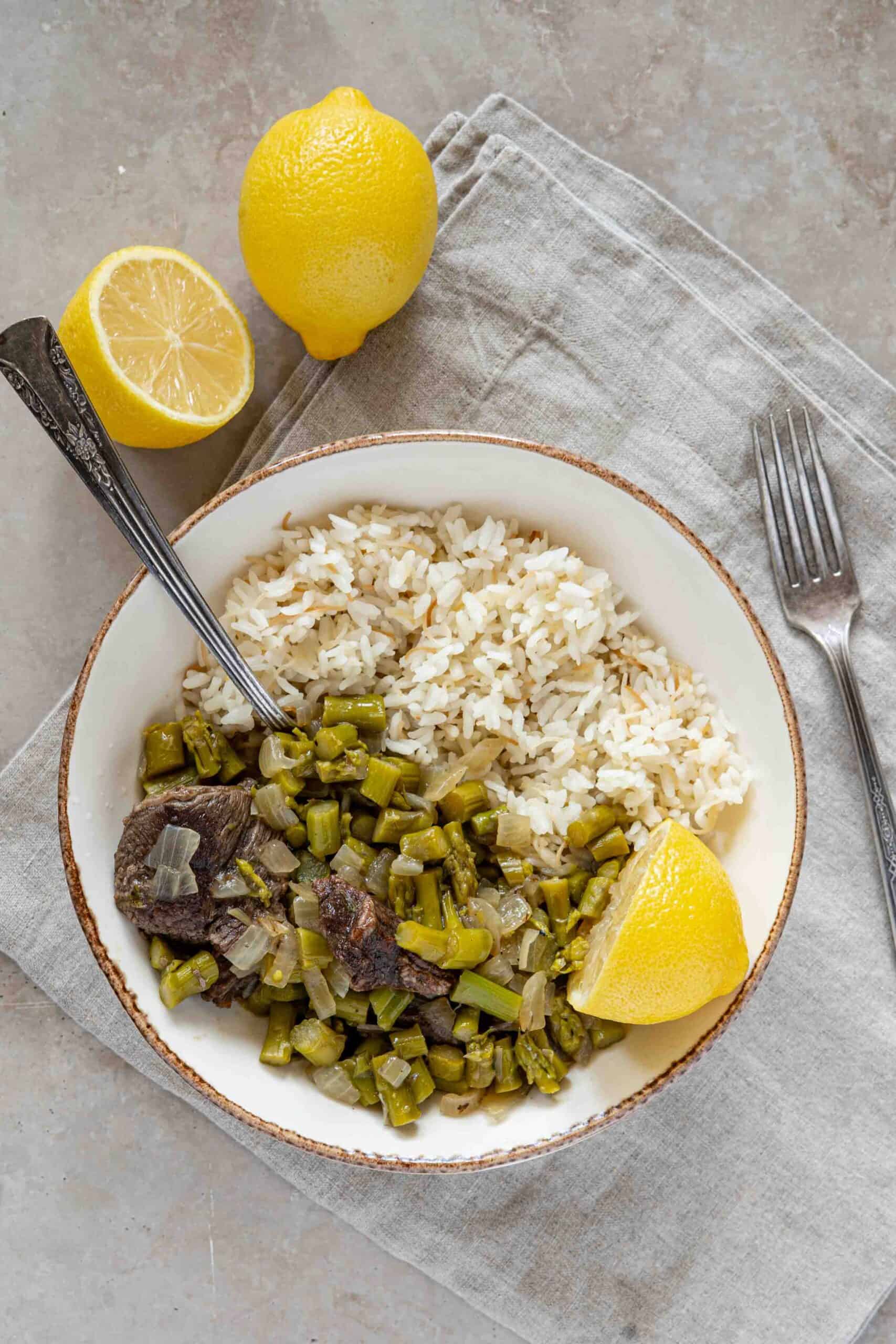 A bowl half cooked asparagus with meat and half vermicelli rice, with fresh lemons ready to squeeze over the stew.