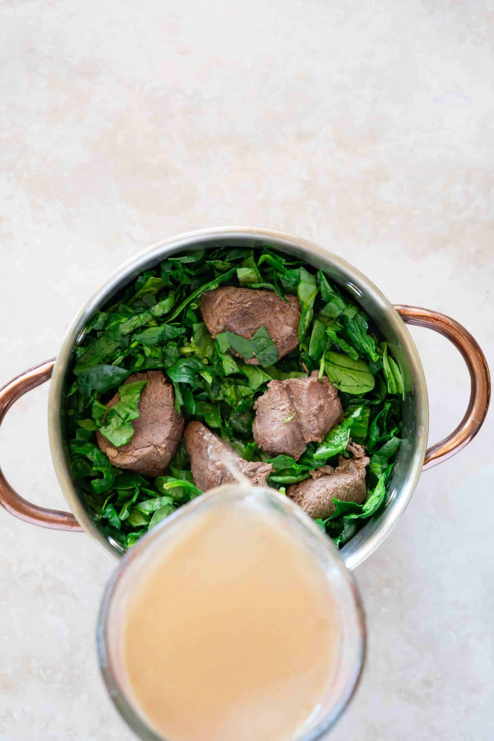 Meat pieces placed over chopped spinach in a pot, then pouring the meat broth evenly over them.