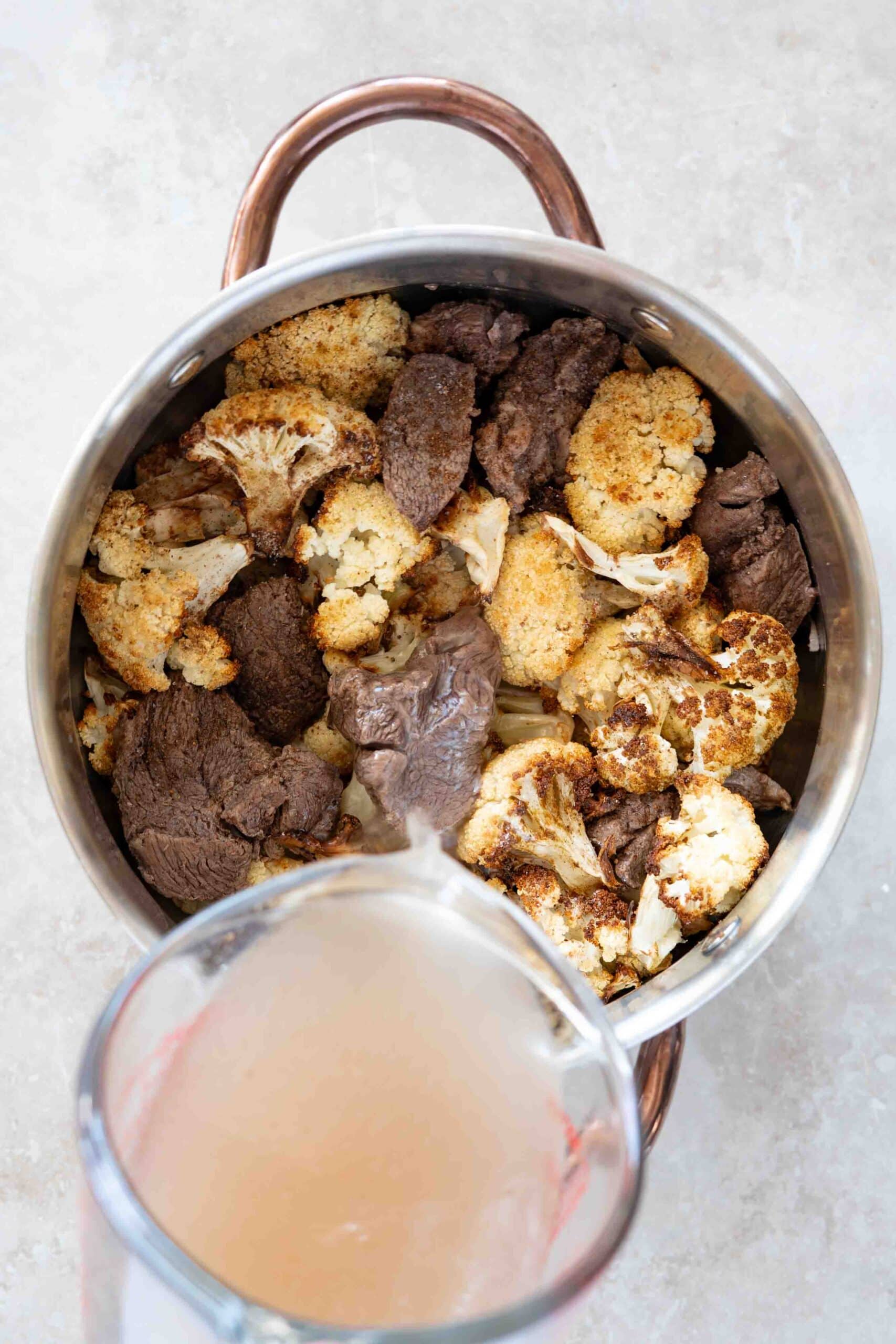 Pouring broth over grilled cauliflowers and lamb chunks in pot.