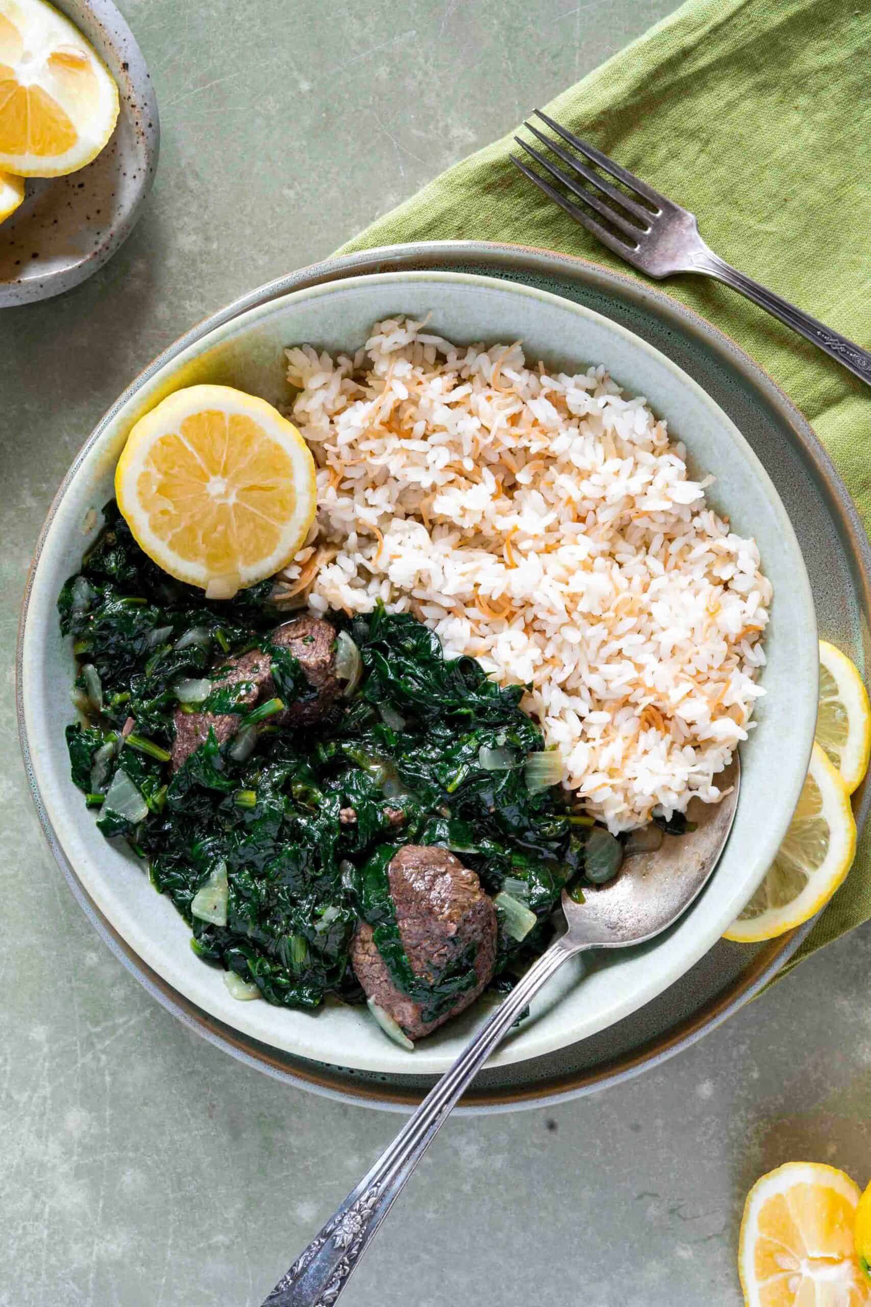 A serving bowl filled with one half spinach stew with lamb pieces and another half of vermicelli rices, with slices of lemon next to it.