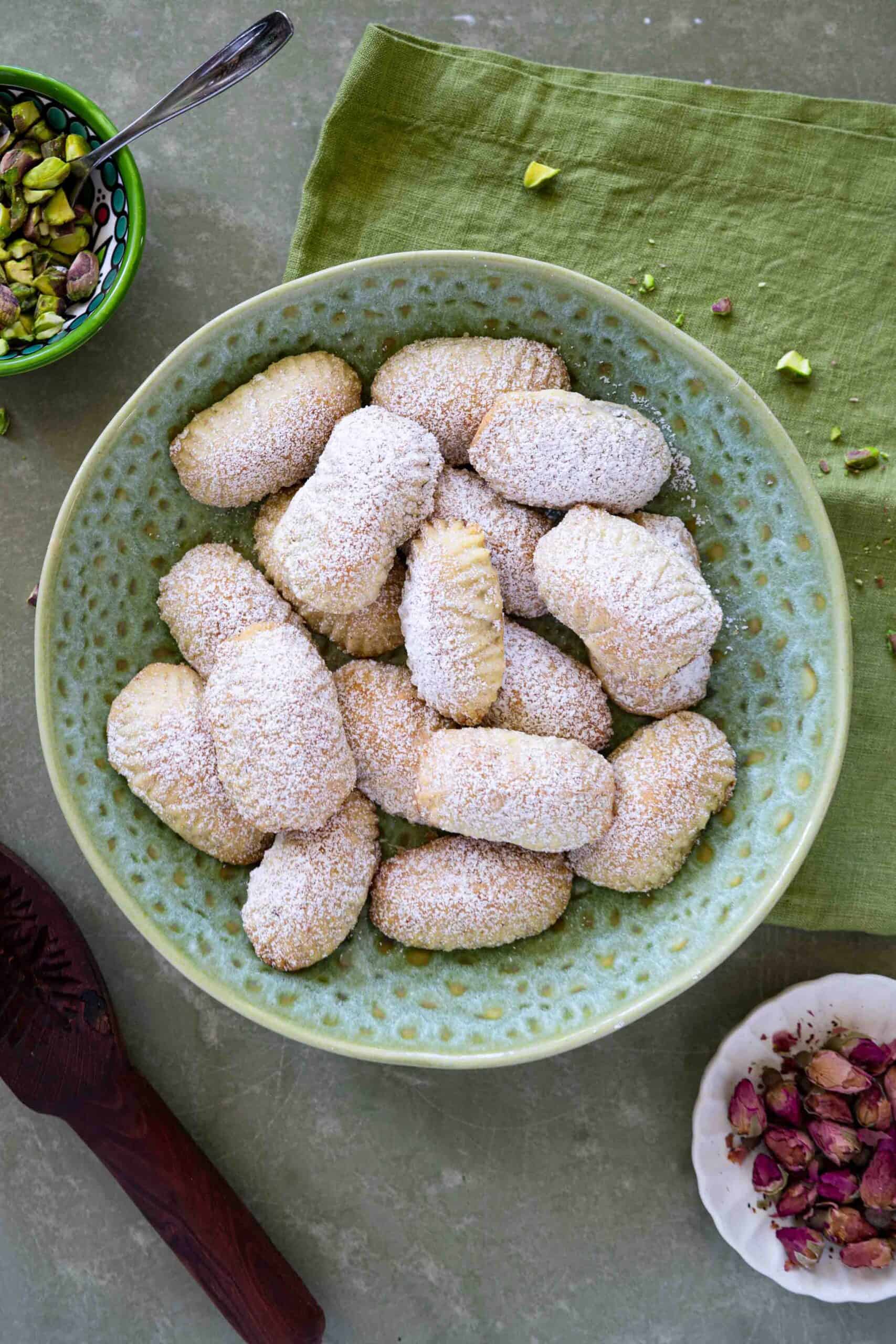 Pistachio maamoul cookies dusted with powdered sugar in a serving bowl. Decorated with dried rose petals. A side of a small plate filled with pistachios and another with hot pink rose petals. With a cookie mold on the side
