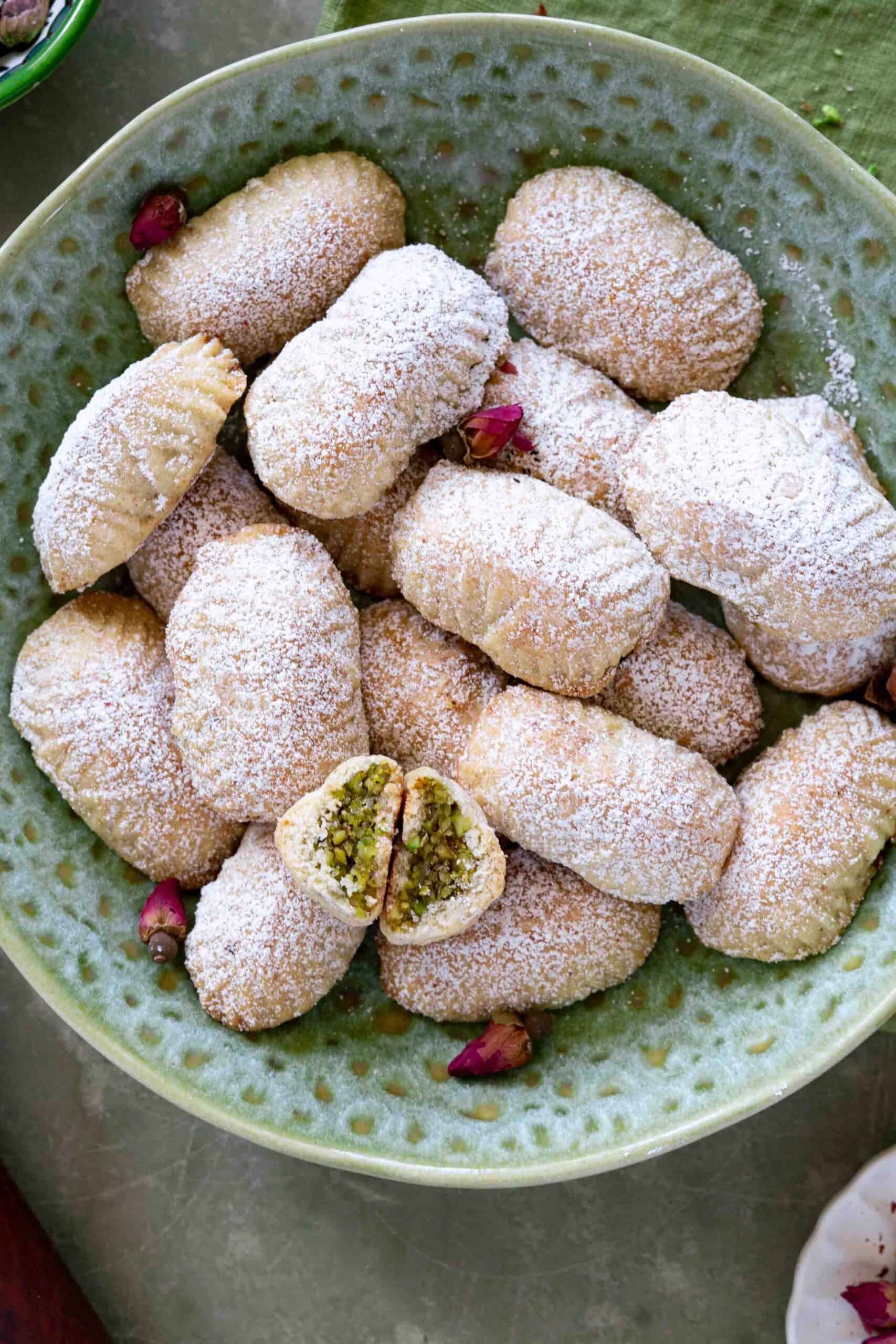 Close up photo of pistachio maamoul cookies dusted with powdered sugar in a bowl. Decorated with dried rose petals.
