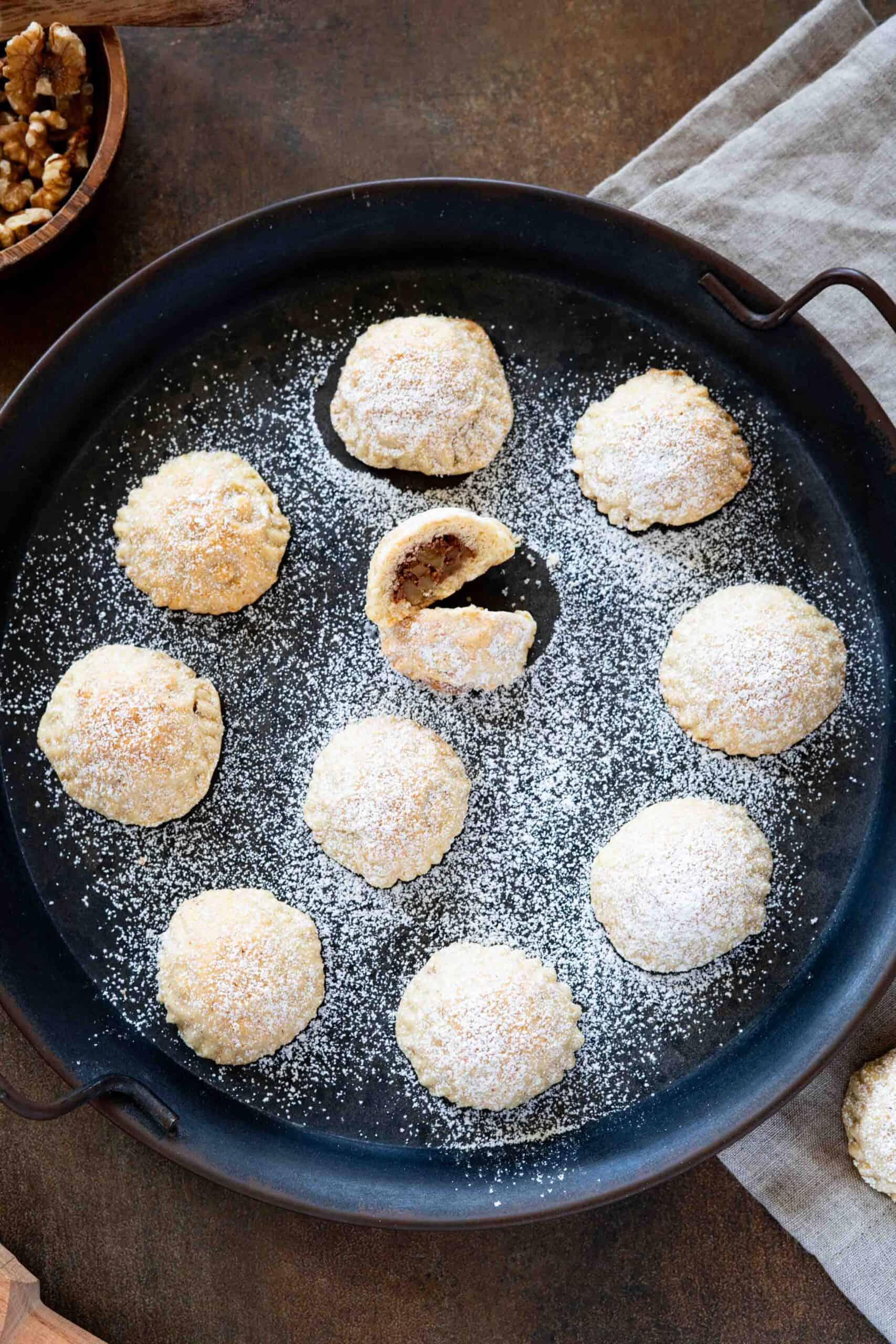 A close-up of a rounded tray filled with walnut maamoul dusted with powdered sugar, with one maamoul split into two pieces to reveal the walnut filling.