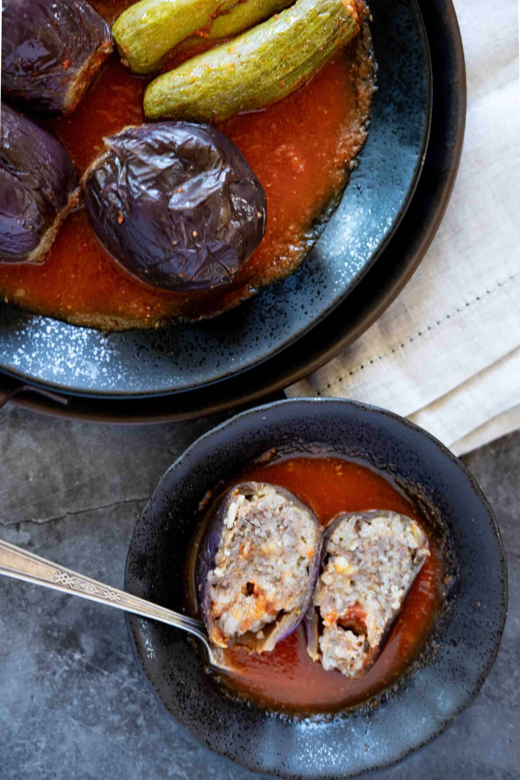 One stuffed eggplant split in half with some tomato sauce, next to a plate with stuffed eggplants and zucchini