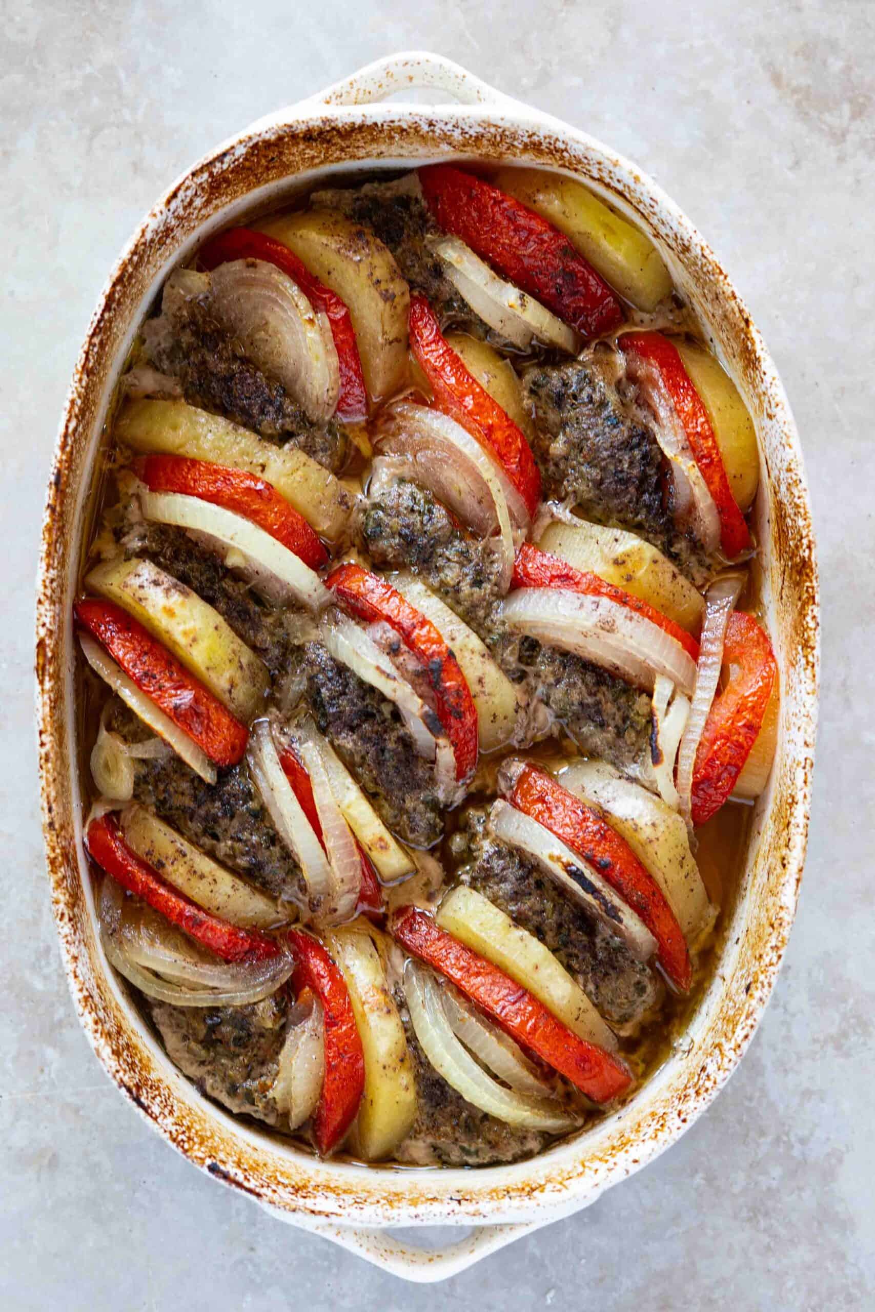 Oval-shaped kafta after baking arranged in a baking dish, with slices of potatoes, tomatoes and onions, placed beside each piece of kafta.