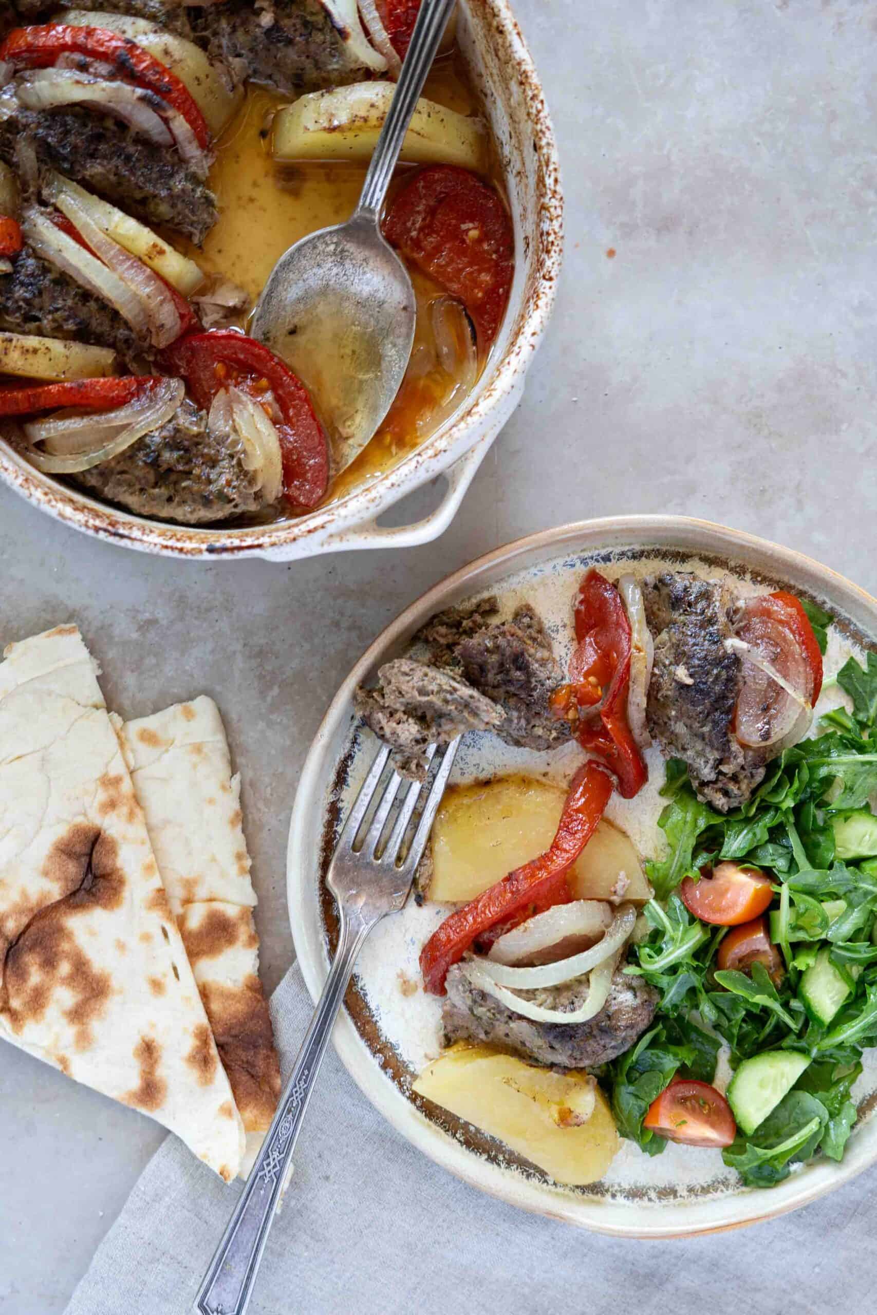 a portion of kafta with potatoes, tomatoes and onions placed in a serving plate next to a side of arugula salad, and a piece of flat bread.