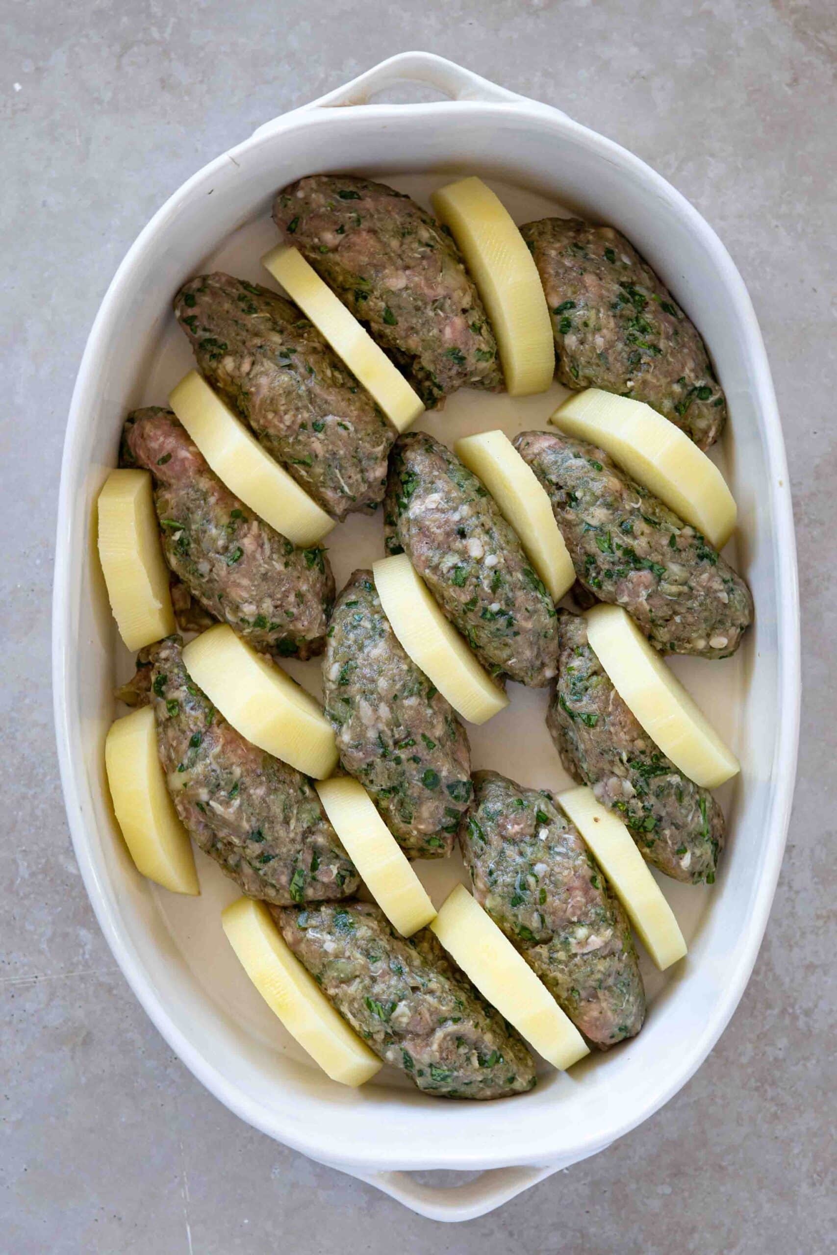 Oval-shaped kafta arranged in a baking dish, with slices of peeled potatoes placed beside each piece of kafta.