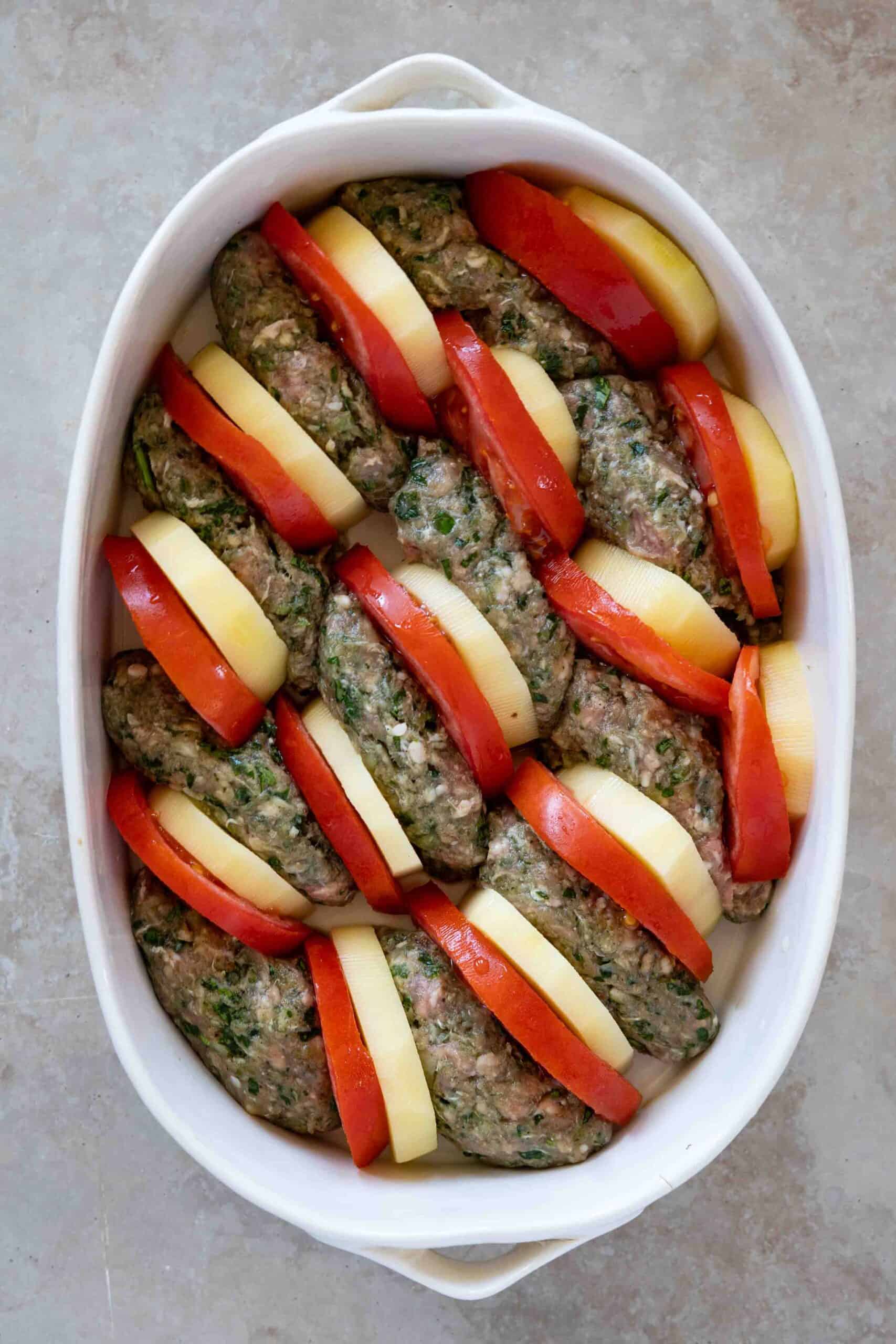 Oval-shaped kafta arranged in a baking dish, with slices of peeled potatoes and tomatoes placed beside each piece of kafta.