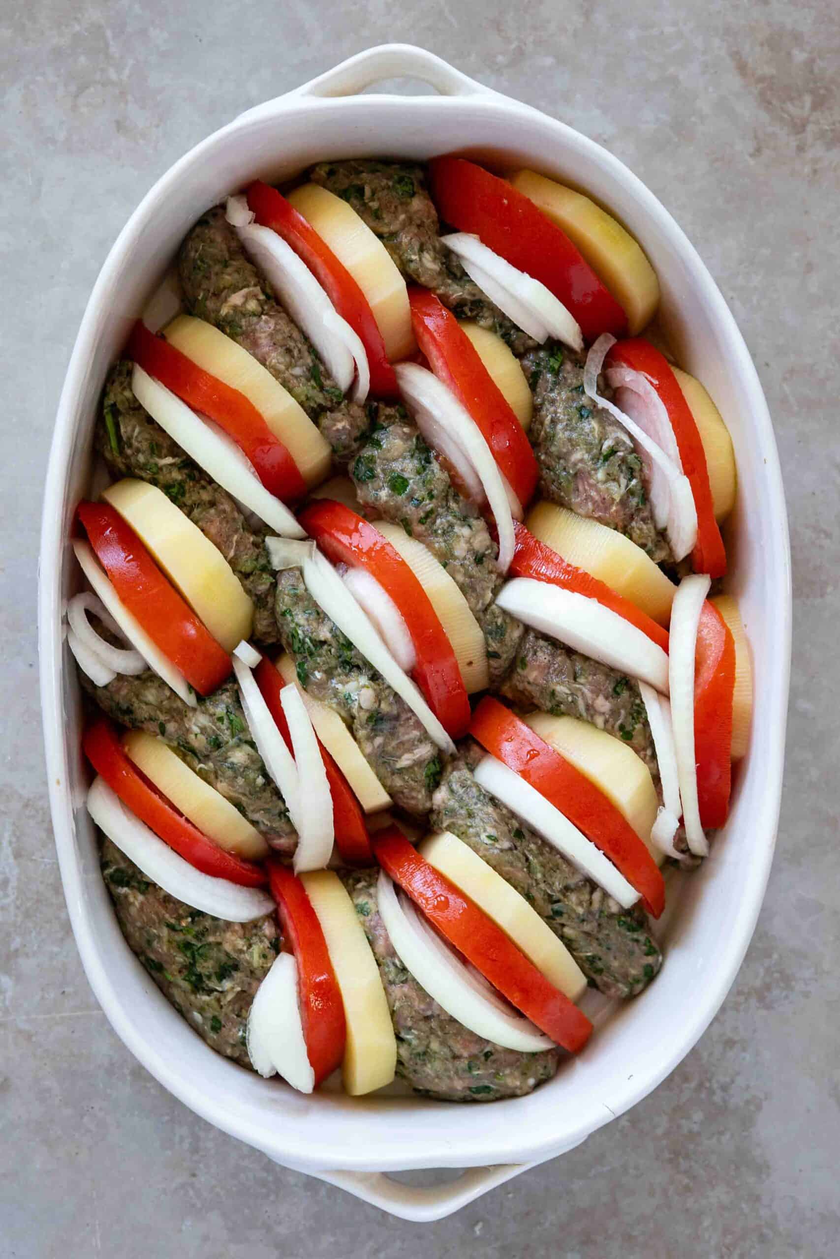 Oval-shaped kafta arranged in a baking dish, with slices of peeled potatoes, and peeled onions and tomatoes placed beside each piece of kafta.