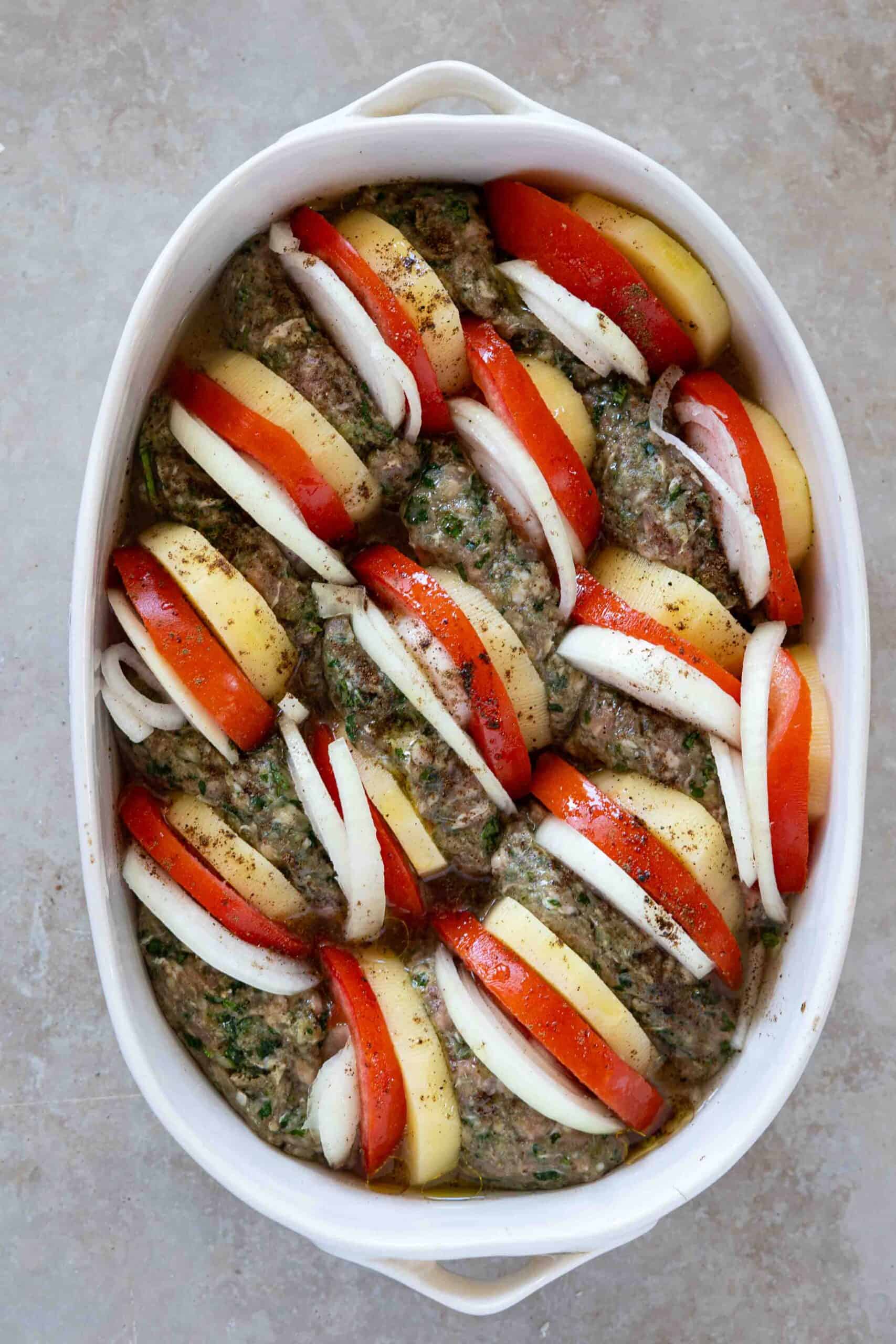 Kafta arranged in a baking dish, with slices of peeled potatoes, and peeled onions and tomatoes placed beside each piece of kafta. Sprinkled with salt and allspice ready for baking.