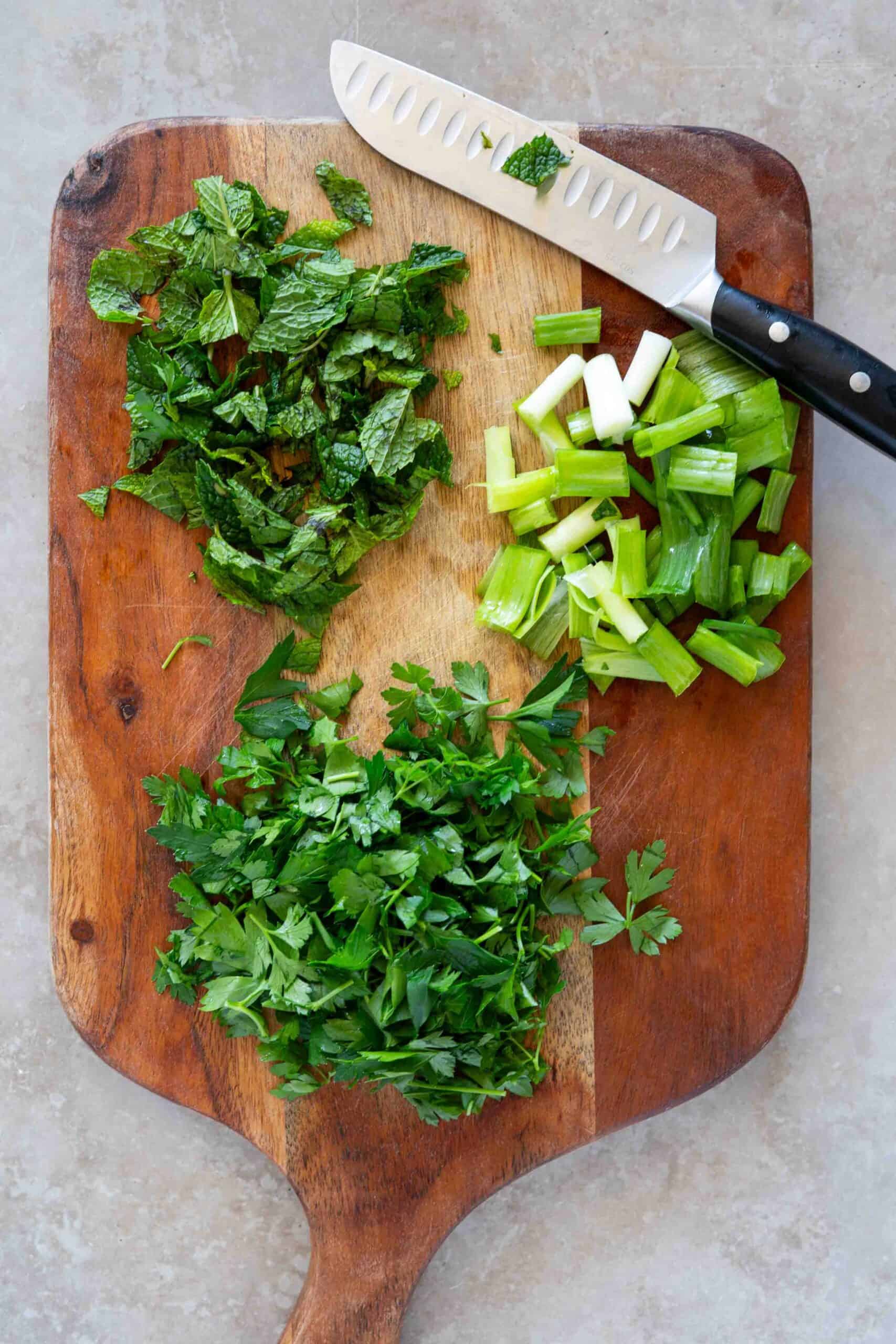 Roughly chopped mint leaves, parsley leaves and green onions on a cutting board.