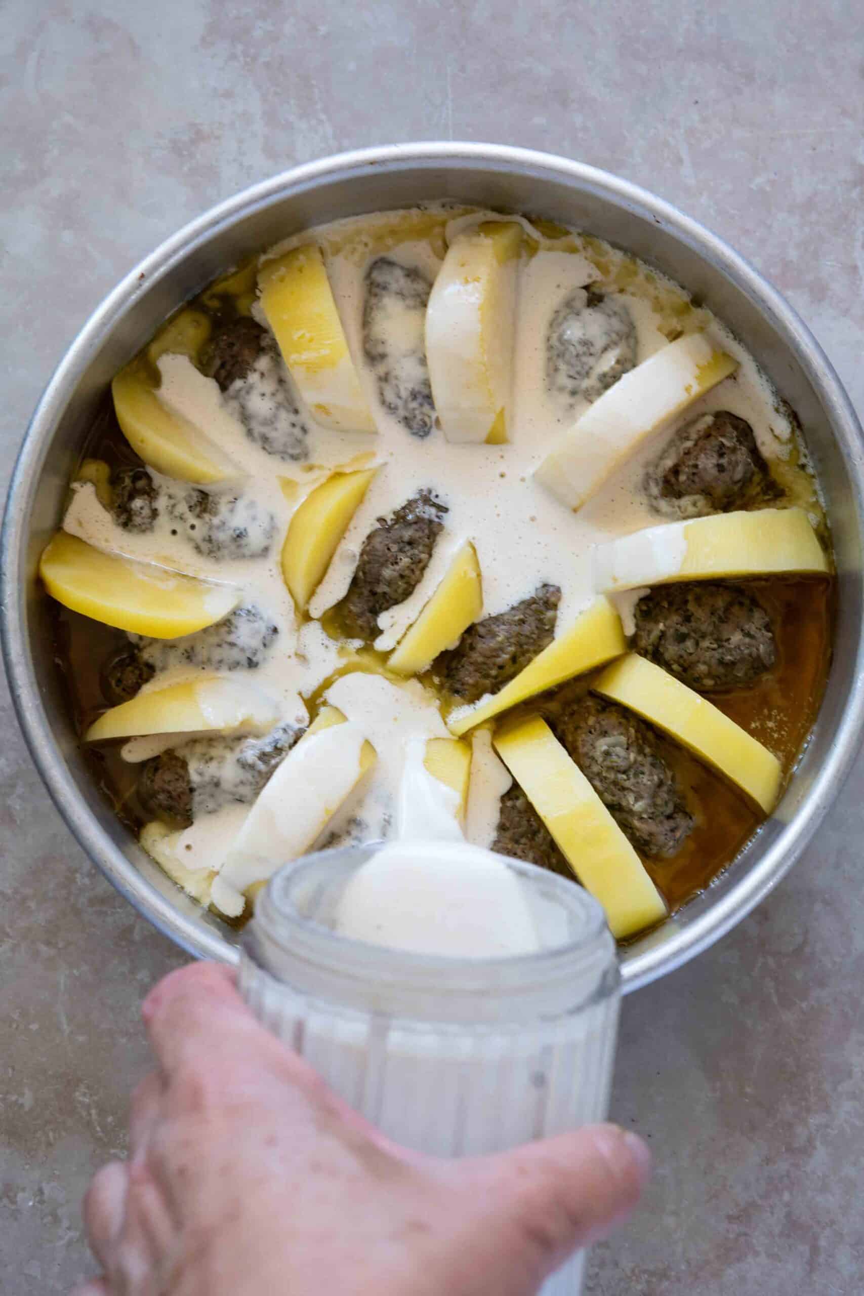Pouring tahini sauce over kafta bil tahini in a baking pot.