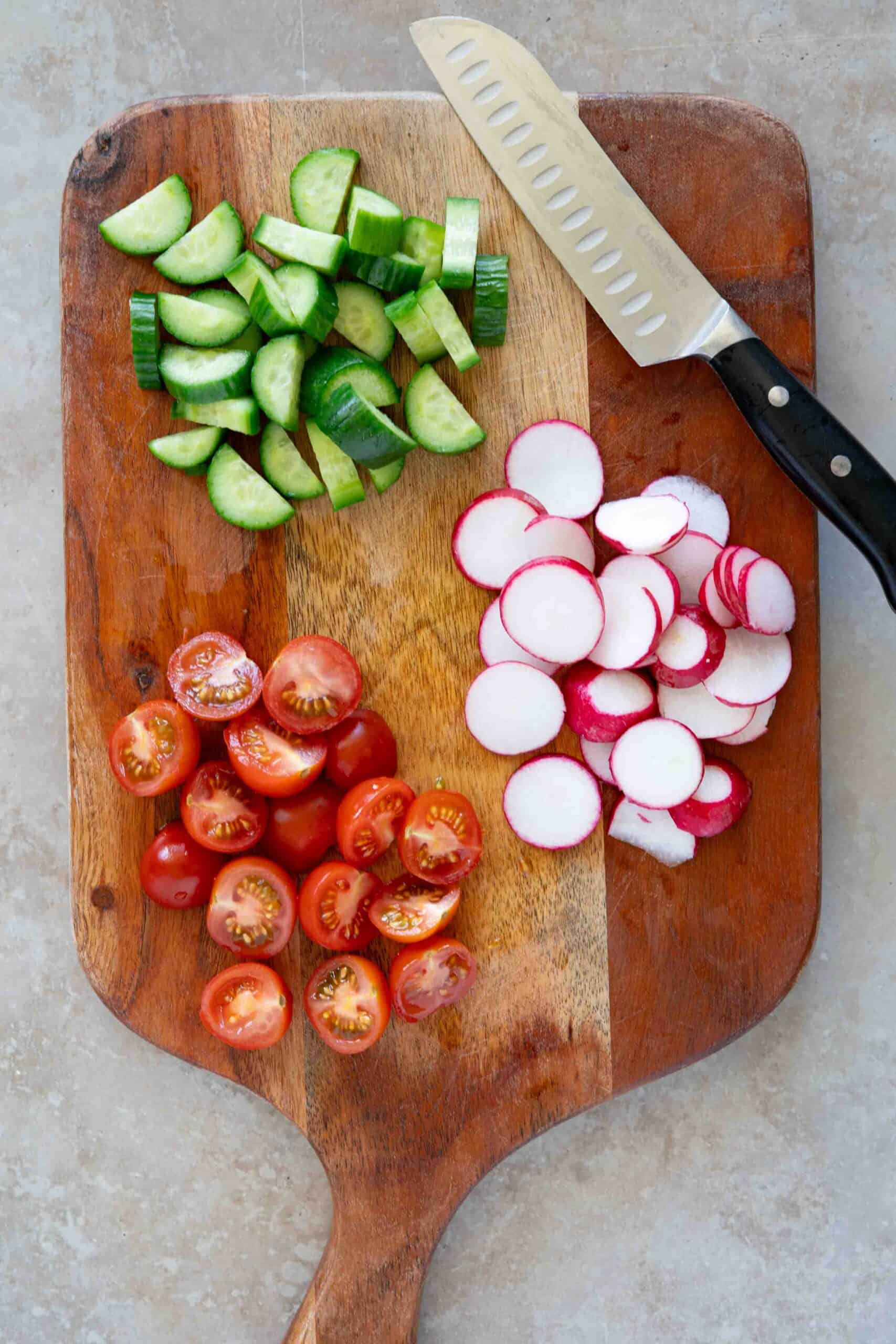 Sliced cucumbers into cubes, cherry tomatoes into halves and sliced radishes on a cutting board.